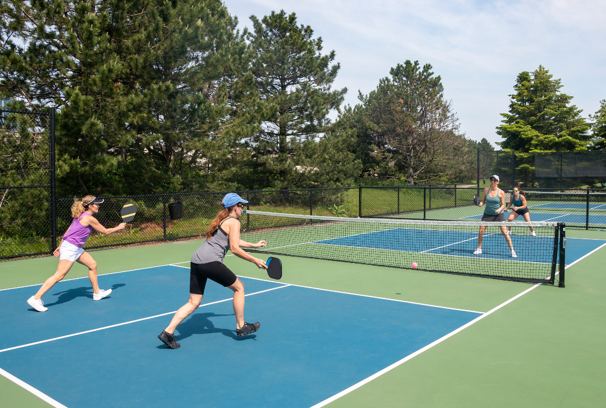 Four women playing pickleball on an outdoor court surrounded by trees.