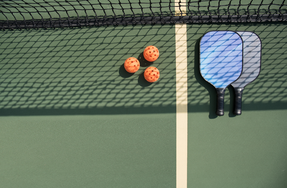 Tennis paddles and three orange tennis balls on a green outdoor tennis court with shadows from the net.