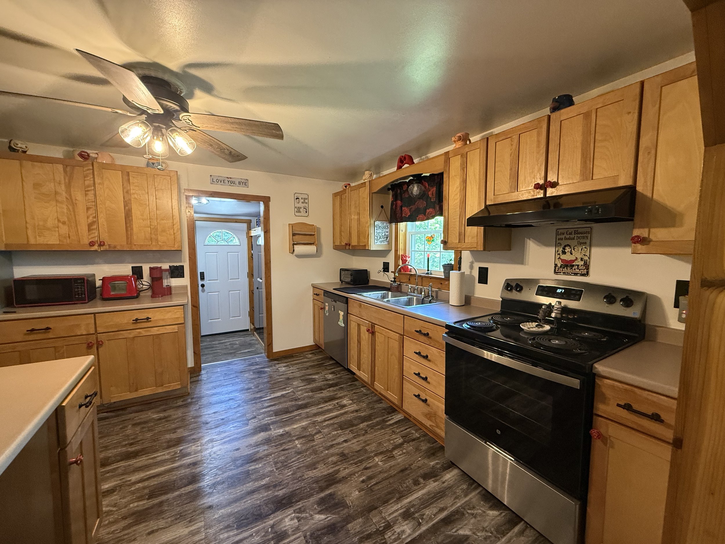 A kitchen with wooden cabinets, a white ceiling fan with lights, a black stove, a microwave, a paper towel roll, a window with a floral curtain, and various decorations and signs on the walls.