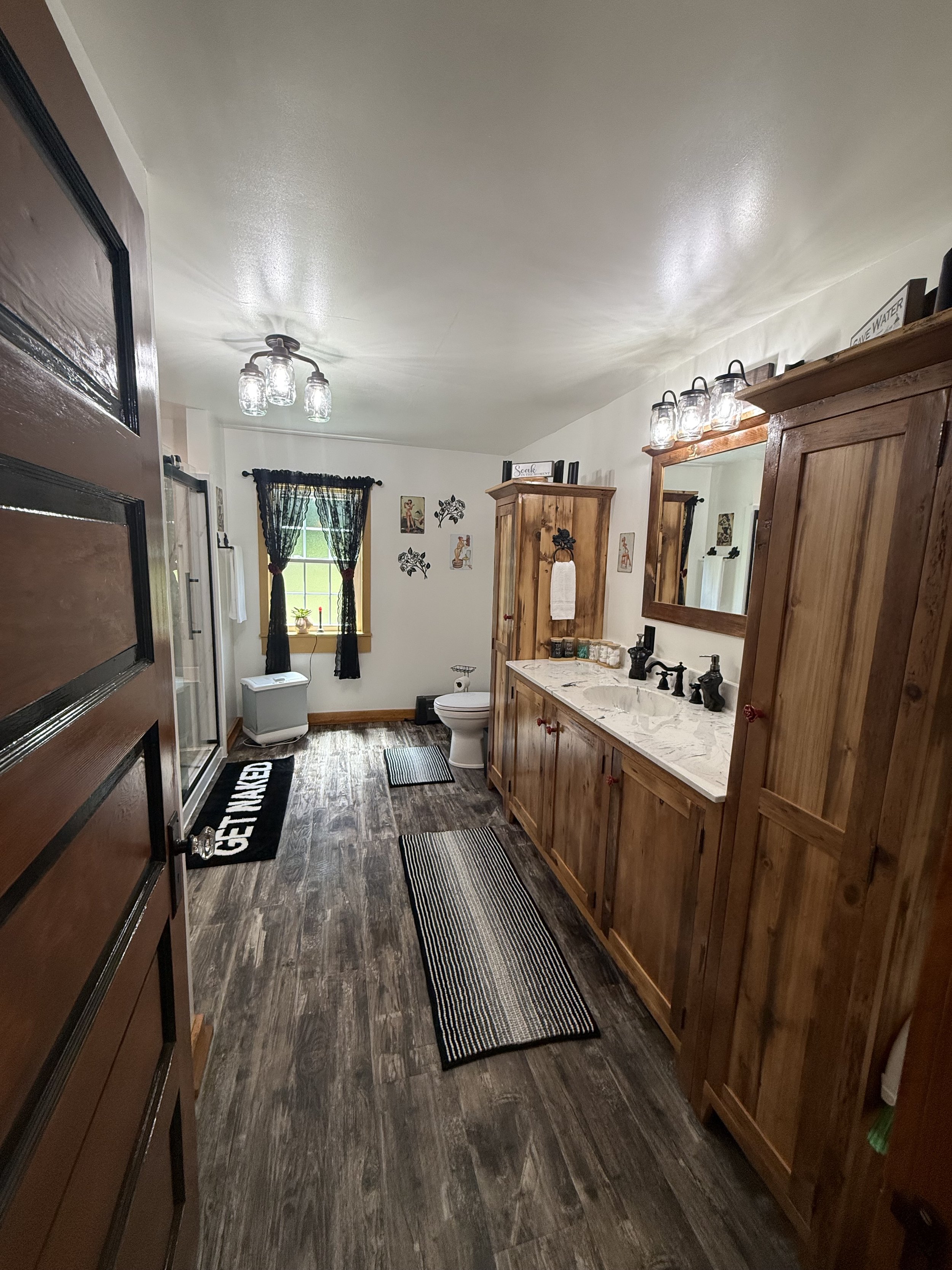 View of a rustic bathroom with wooden cabinets, a marble countertop, a mirror, a toilet, a window with black curtains, and a ceiling light fixture. There are striped rugs on the floor and decorative wall art.