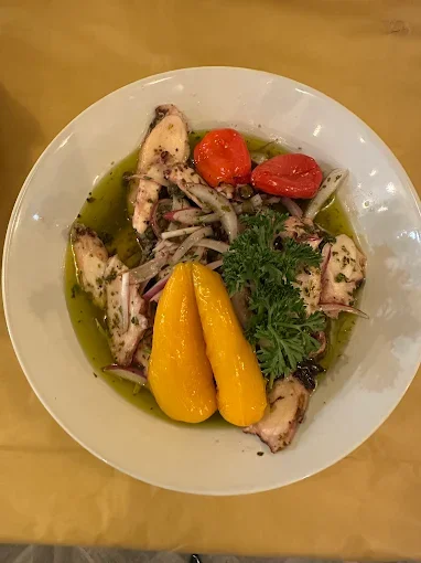 Salad with grilled octopus, cherry tomatoes, yellow peppers, red onion, parsley, and green oil dressing in a white bowl.