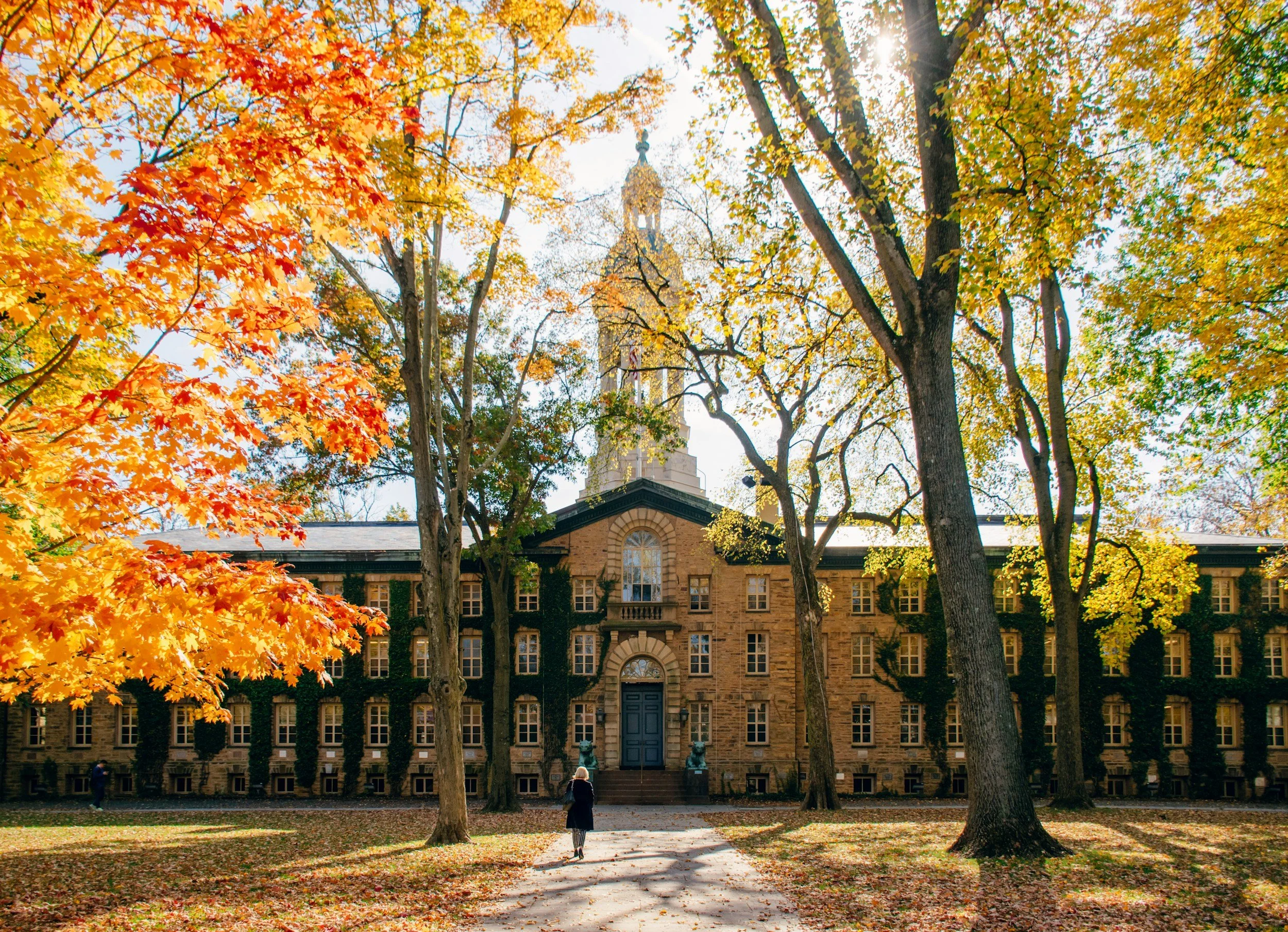 A historic brick building surrounded by large trees with colorful fall leaves, some people walking on the path in front of the building, and sunlight filtering through the trees. Ivy League Schools in the Fall.