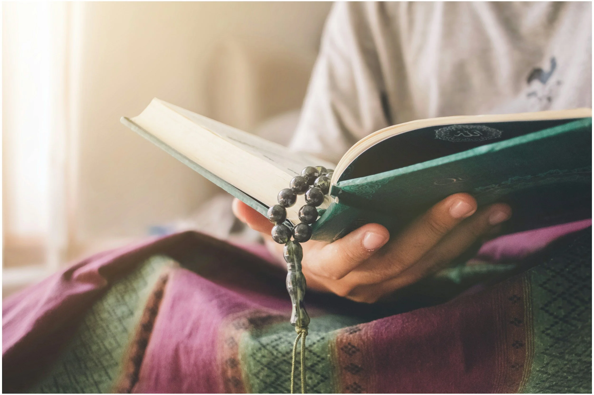Person holding a small book or notepad with a bracelet of dark beads around their wrist, wearing a patterned shirt and pink jacket, with a blurred indoor background.

Combining Education with Islam