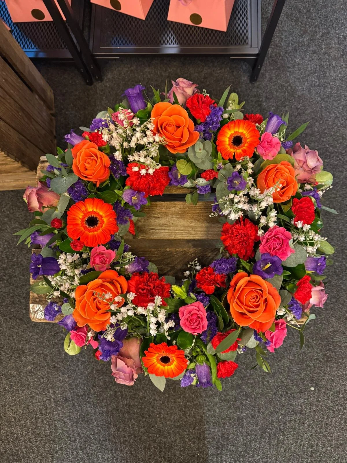 A colorful flower arrangement in a circular wreath shape, featuring orange roses, red carnations, pink roses, purple bellflowers, orange and black gerbera daisies, white baby's breath, and various green foliage. The arrangement is placed on a wooden surface with a dark carpeted floor underneath.