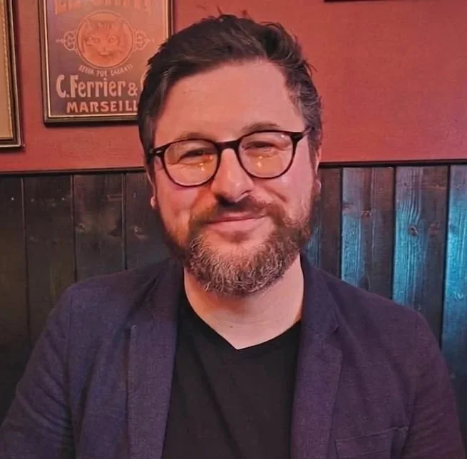 A smiling man with glasses, a beard, and short hair, sitting in a dimly lit restaurant or bar with wooden paneling and framed posters on the wall behind him.