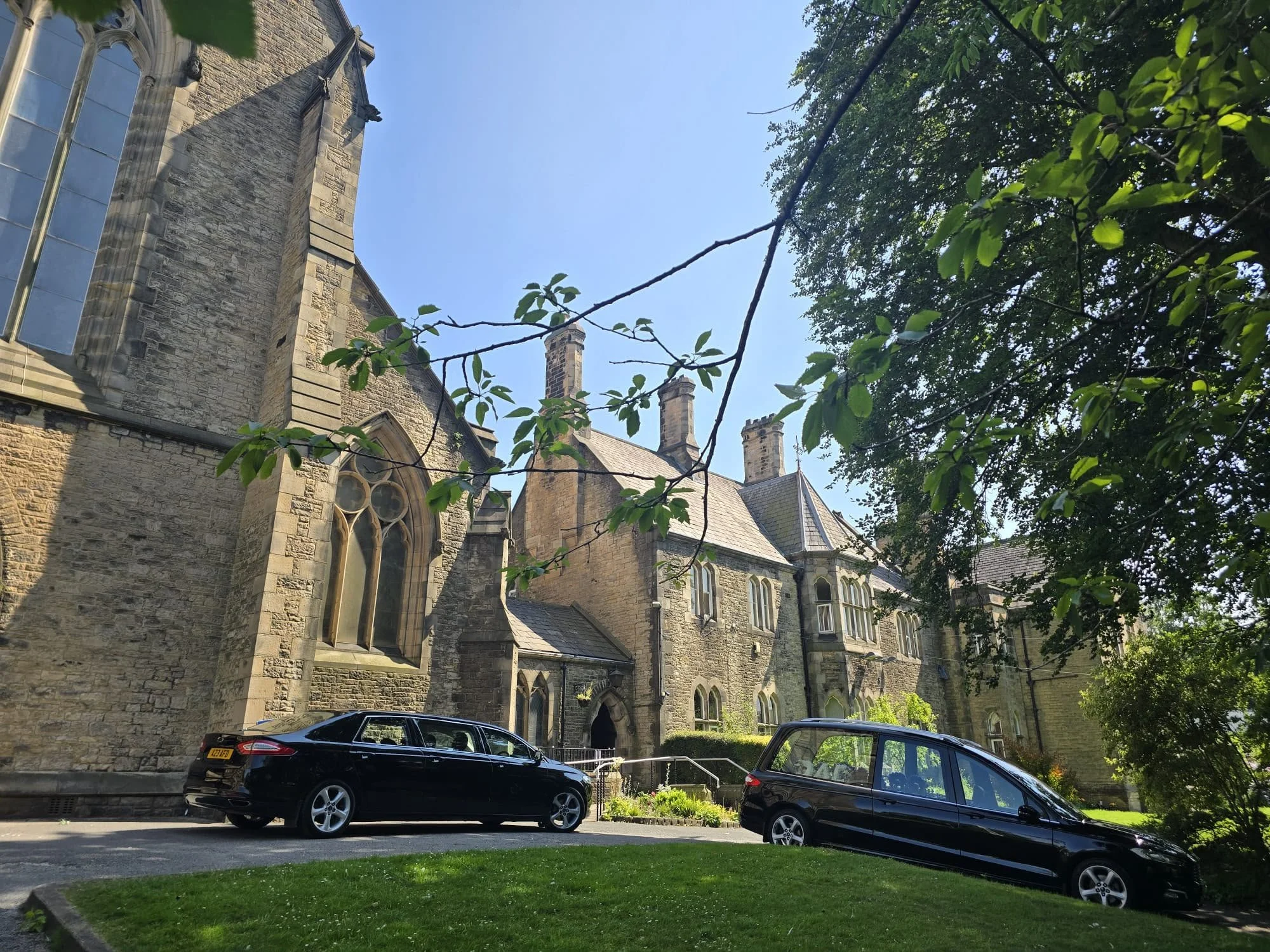 A stone church with gothic windows and multiple chimneys, surrounded by green trees, with cars parked in front, on a sunny day.