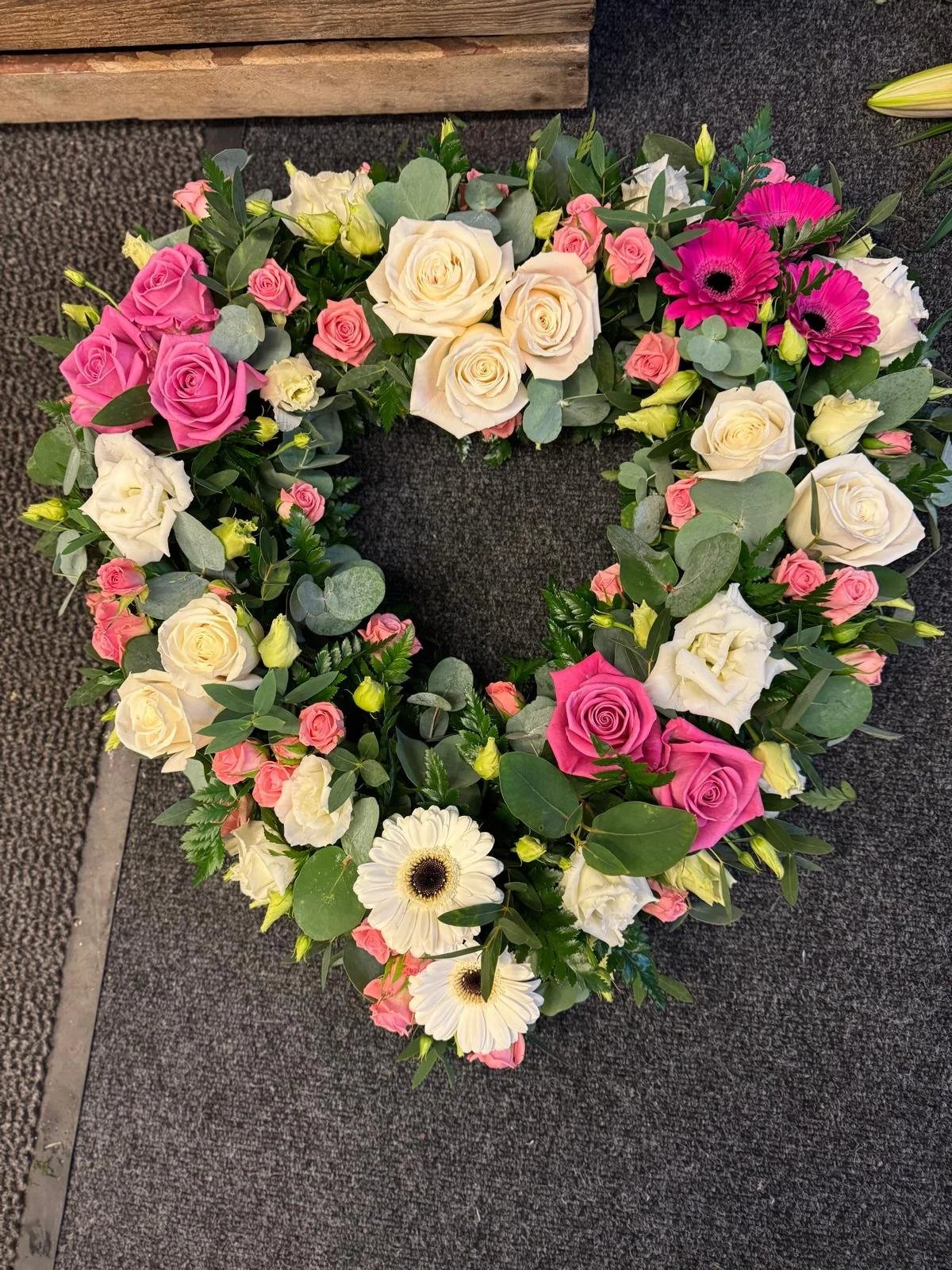A floral heart-shaped arrangement with pink, white, and cream roses, white gerbera daisies, and green foliage on black and gray flooring.