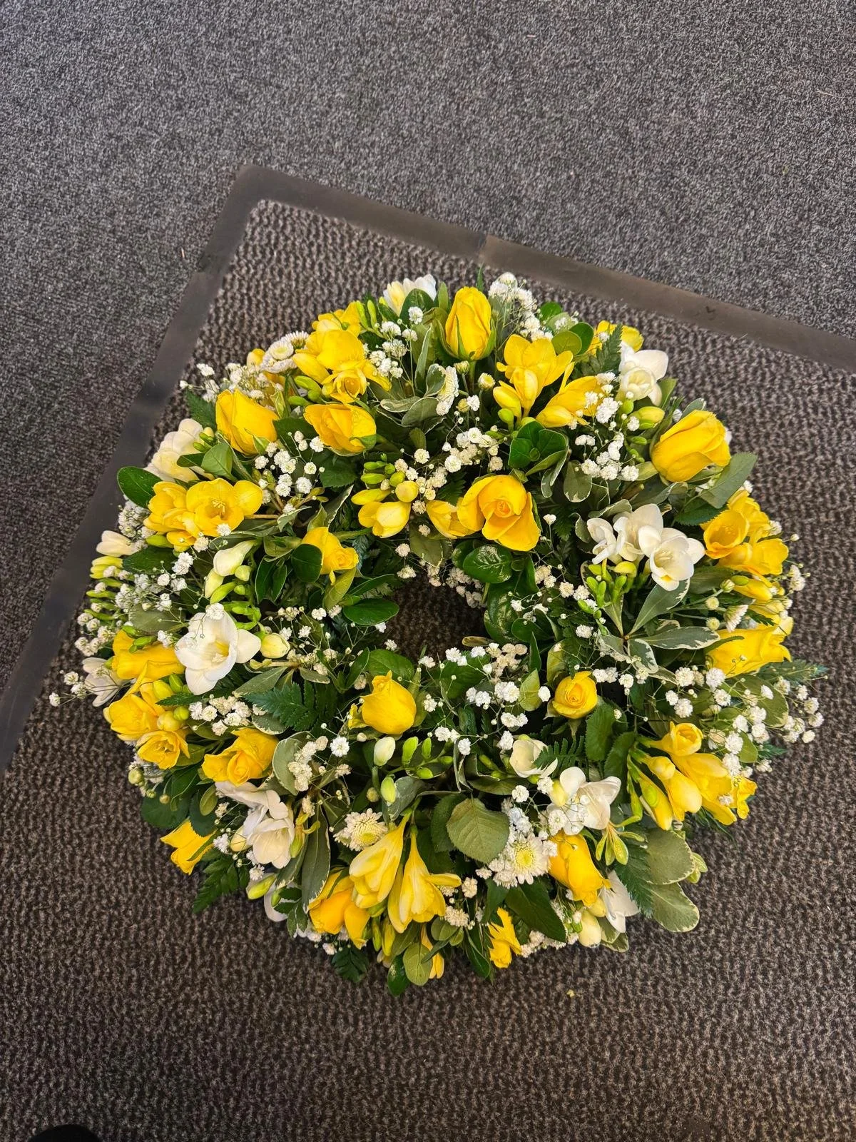 A floral wreath made of yellow and white flowers, with green leaves, placed on a gray textured carpet.