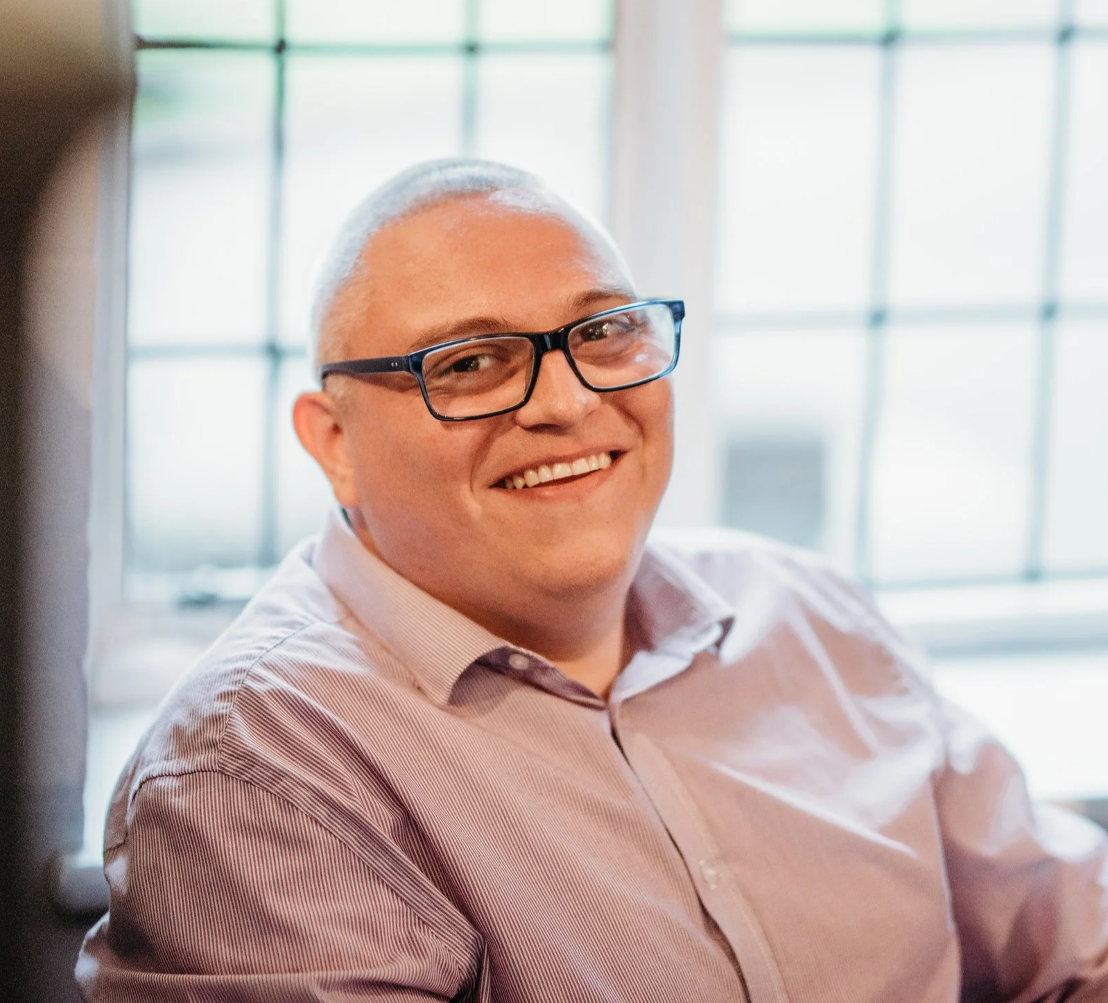 A smiling man with glasses sitting near a window in a well-lit room.