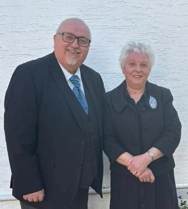 A smiling man and woman standing side by side against a white brick wall. The man is wearing a black suit with a striped tie and glasses, and the woman is dressed in dark clothing with a badge on her jacket.
