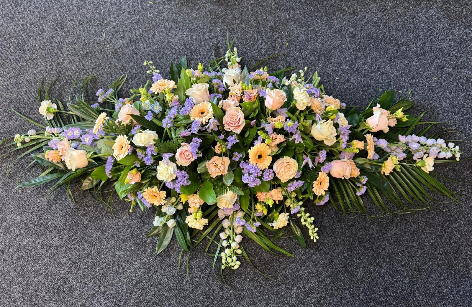 A large floral arrangement with white and peach roses, purple and peach gerbera daisies, lavender, and green foliage, laying on a gray carpet.