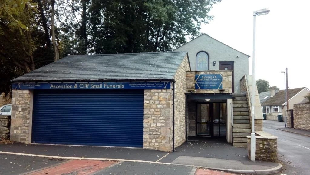 A small funeral home with a stone exterior, blue garage door, and stairs leading to an upper entrance with a glass door, situated on a quiet residential street.