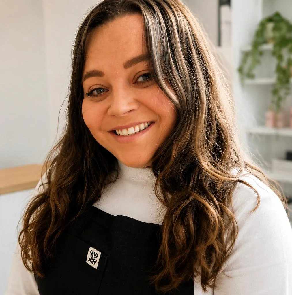 A smiling woman with long, wavy brown hair, wearing a black apron over a white shirt, standing in a bright room with shelves and green plants in the background.