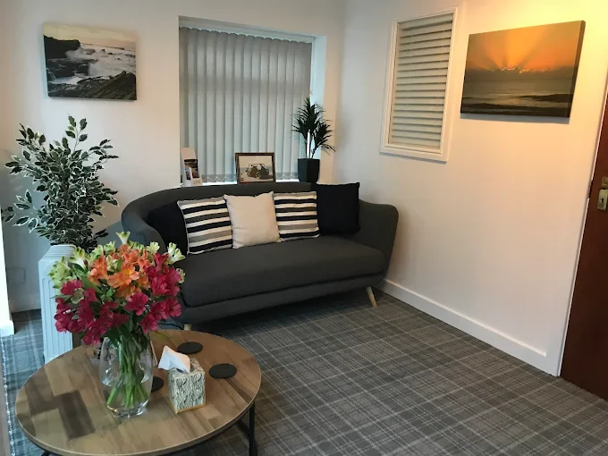 A cozy living room with a dark gray sofa accented with white and black striped pillows, a wooden coffee table with a vase of pink and orange flowers, framed photos, and decorative plants, with wall art depicting sunsets and a window with vertical blinds.
