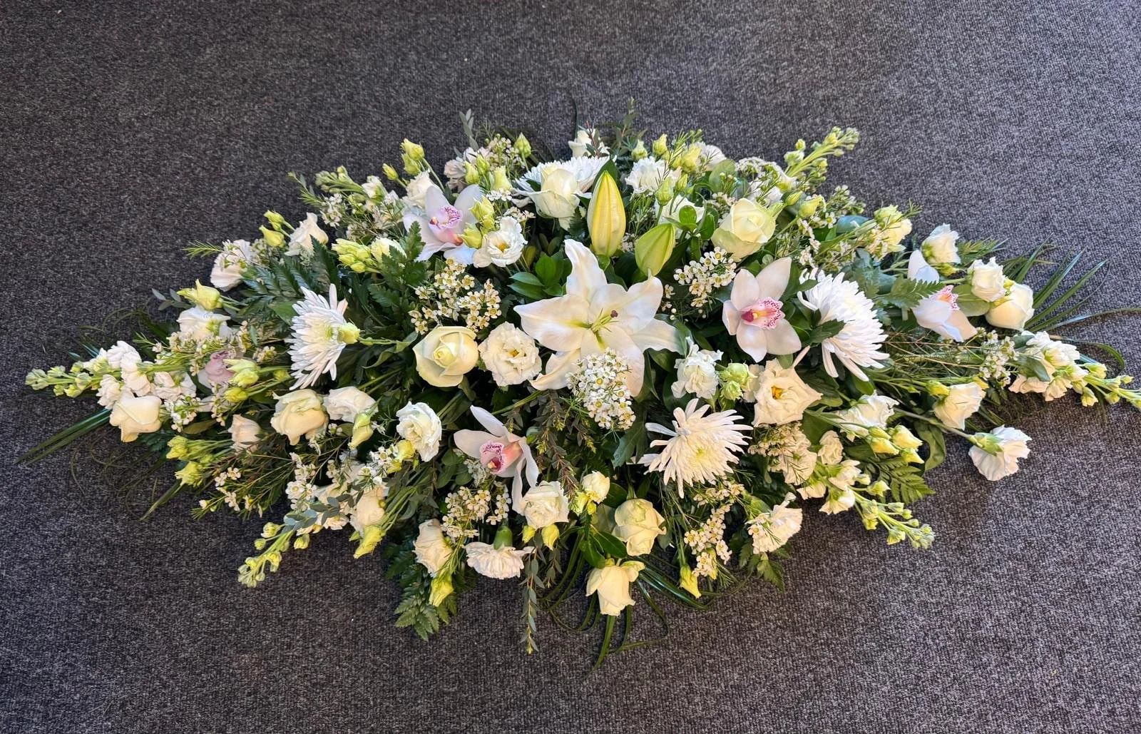 A large white floral arrangement on a gray carpeted surface, featuring white lilies, roses, daisies, orchids, and various greenery.