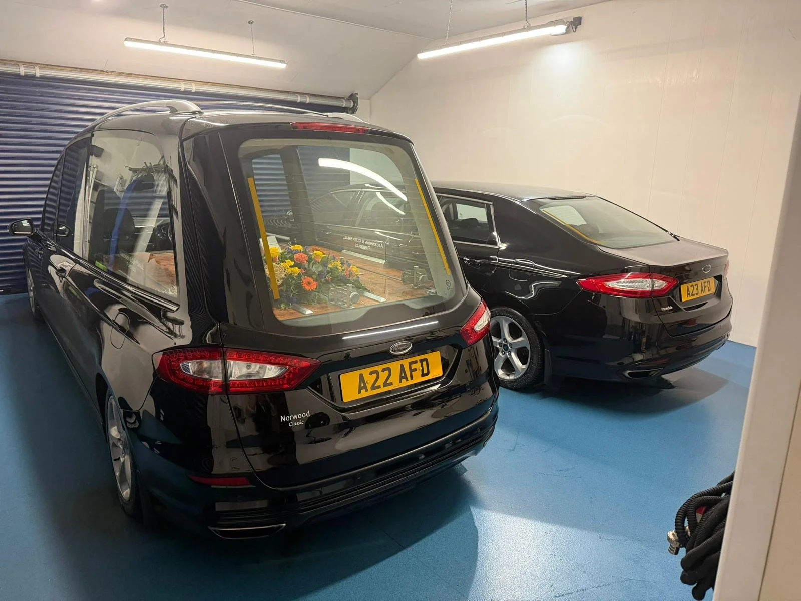 Two black funeral cars parked inside a garage, one with a casket and flowers inside.