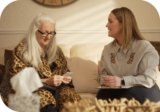 An elderly woman with long white hair and glasses, wearing a leopard print robe, is sitting on a beige couch, looking at a photograph. Next to her, a younger woman with shoulder-length brown hair, wearing a light gray shirt with embroidered flowers, is sitting and smiling at her. There is a tissue box on a table in the foreground and a large clock on the wall behind them.