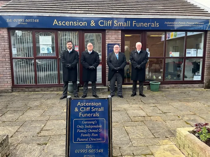 Four men in suits standing outside a funeral home named 'Ascension & Cliff Small Funerals'. There is a blue signboard with the funeral home details in front of them. The building has large windows and a brick exterior.