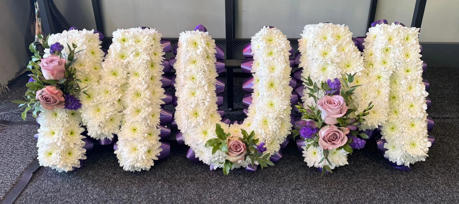 Large floral arrangement spelling out the word 'MOM' with white flowers, accented with pink roses, purple flowers, and greenery, decorated with purple ribbons on a dark carpeted floor.
