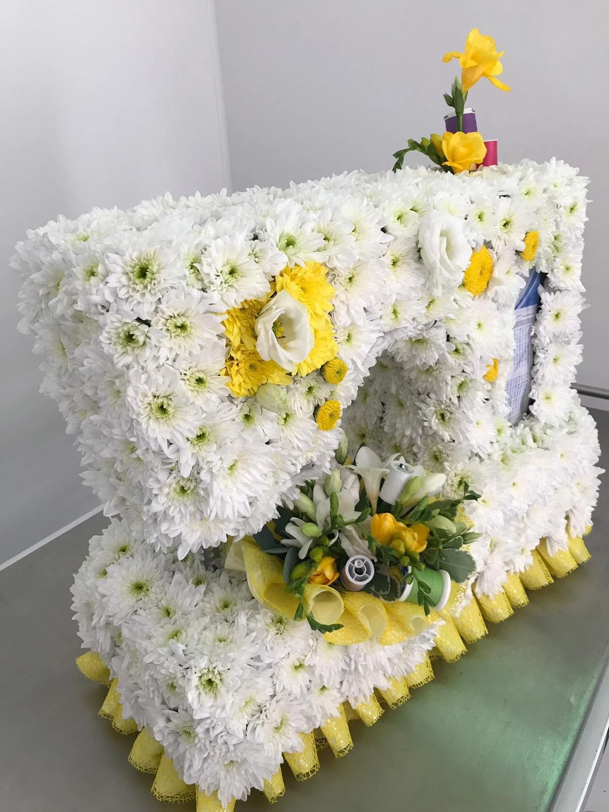 Large floral arrangement made of white flowers with yellow accents, resembling a coffin or casket, decorated with flowers and medical supplies.