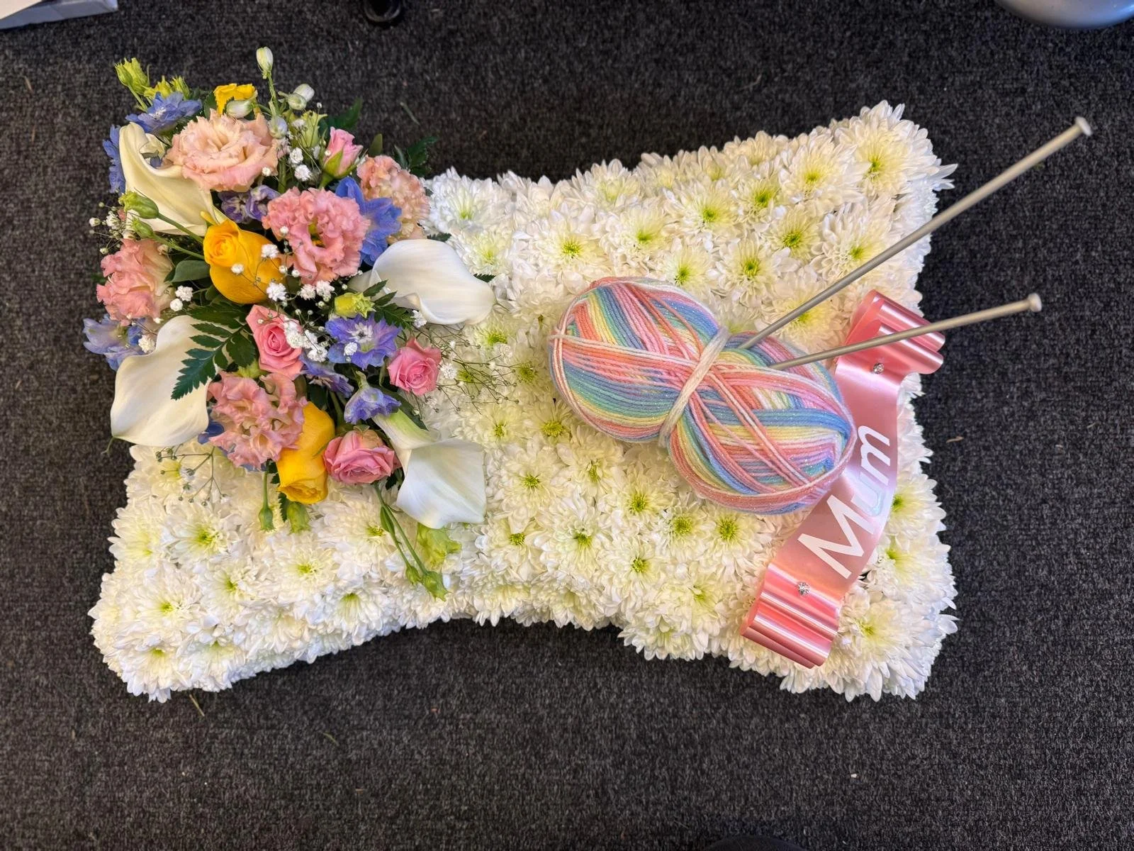 A floral arrangement of mixed flowers including roses, calla lilies, and other blooms on a flower-covered surface with a ball of pastel-colored yarn and knitting needles.