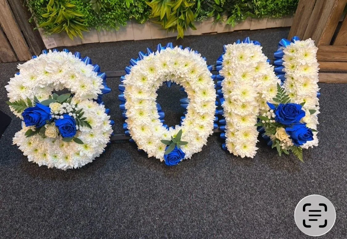 Flower arrangement spelling 'LOVE' with white flowers, accented by blue roses and greenery, on a dark carpeted surface.