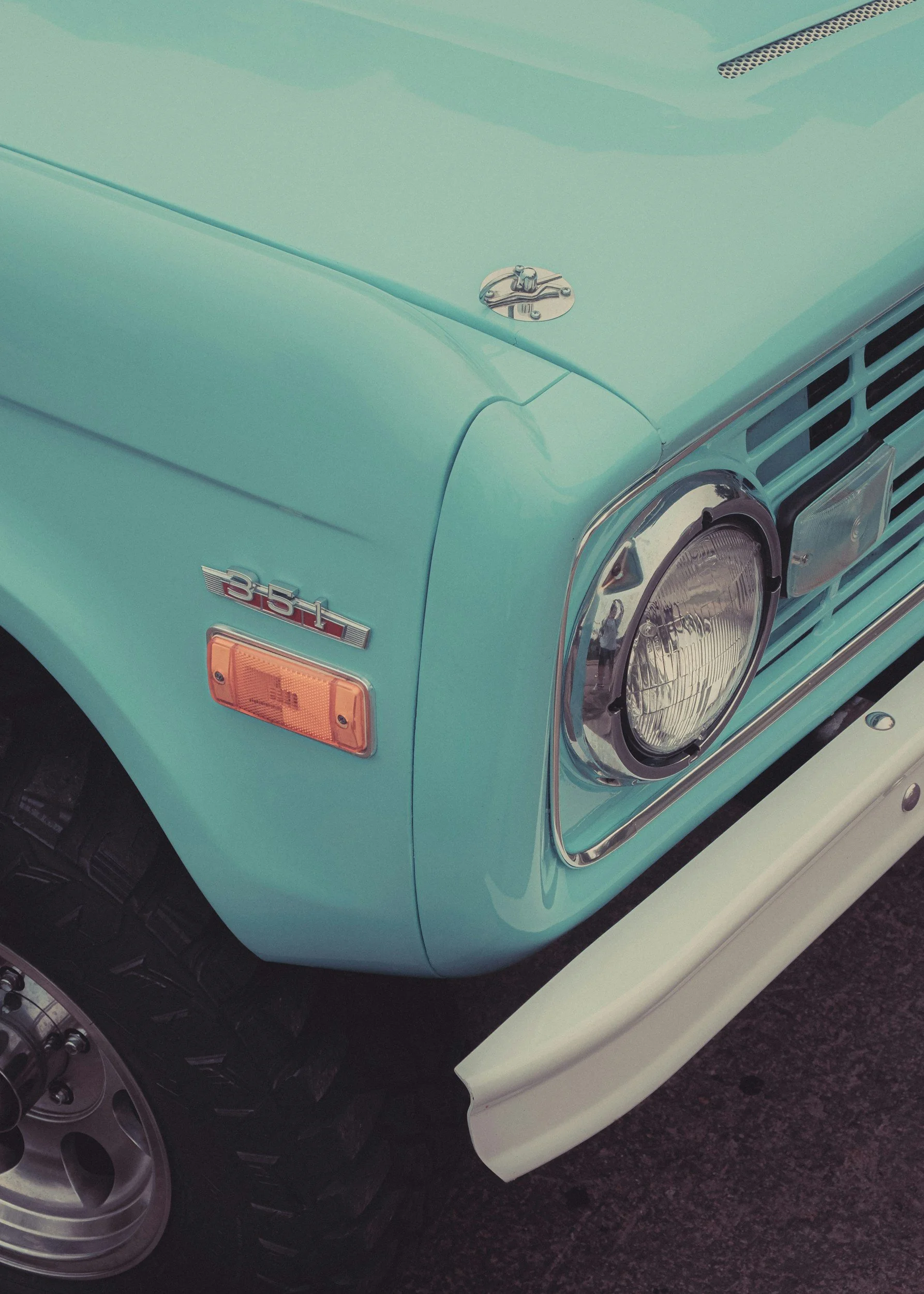 Close-up of the front part of a light blue vintage car, showing the headlight, turn signal, part of the hood with a chrome emblem, and a white bumper.
