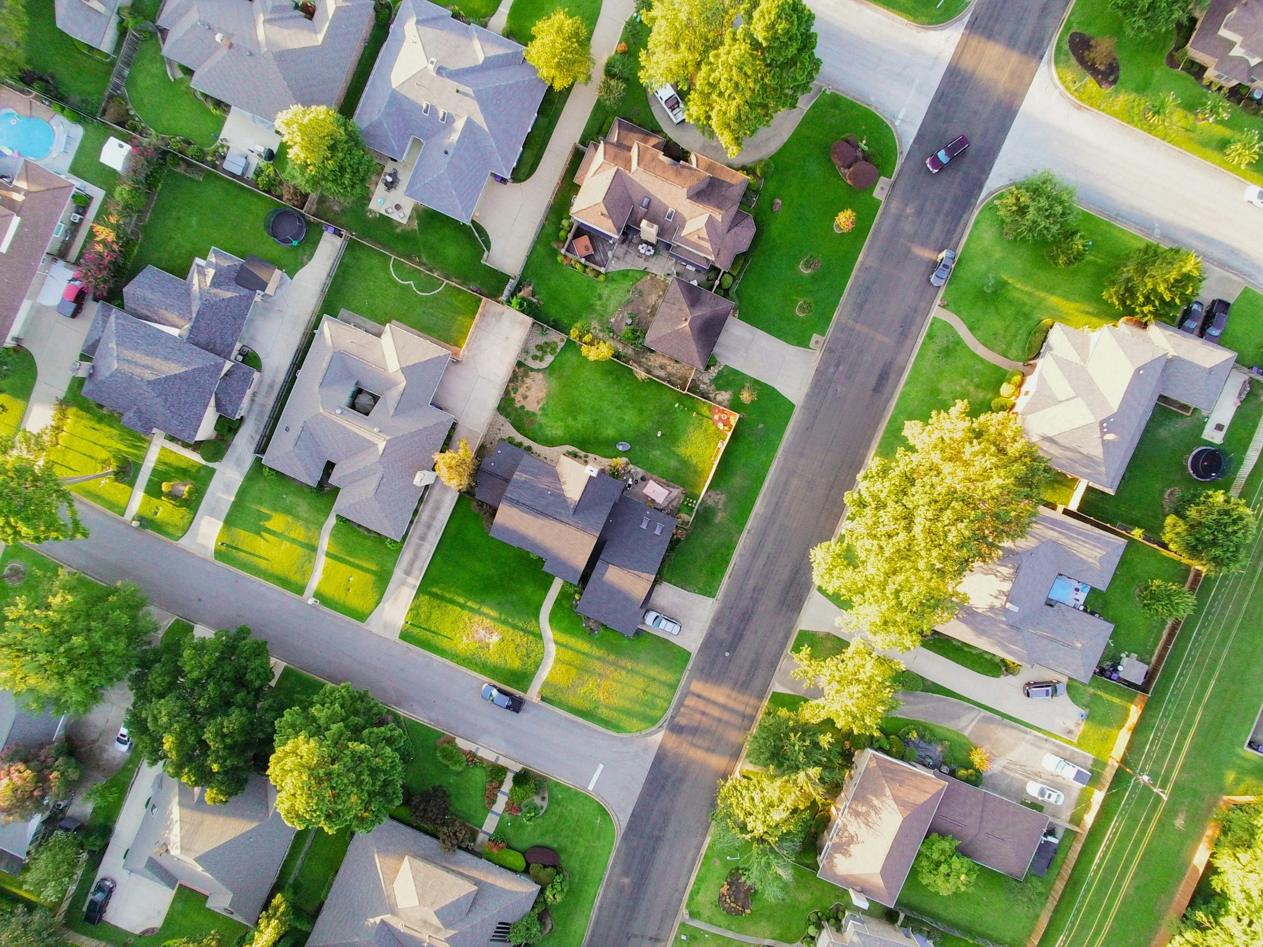 Aerial view of a suburban neighborhood with houses, lawns, trees, sidewalks, and parked cars.