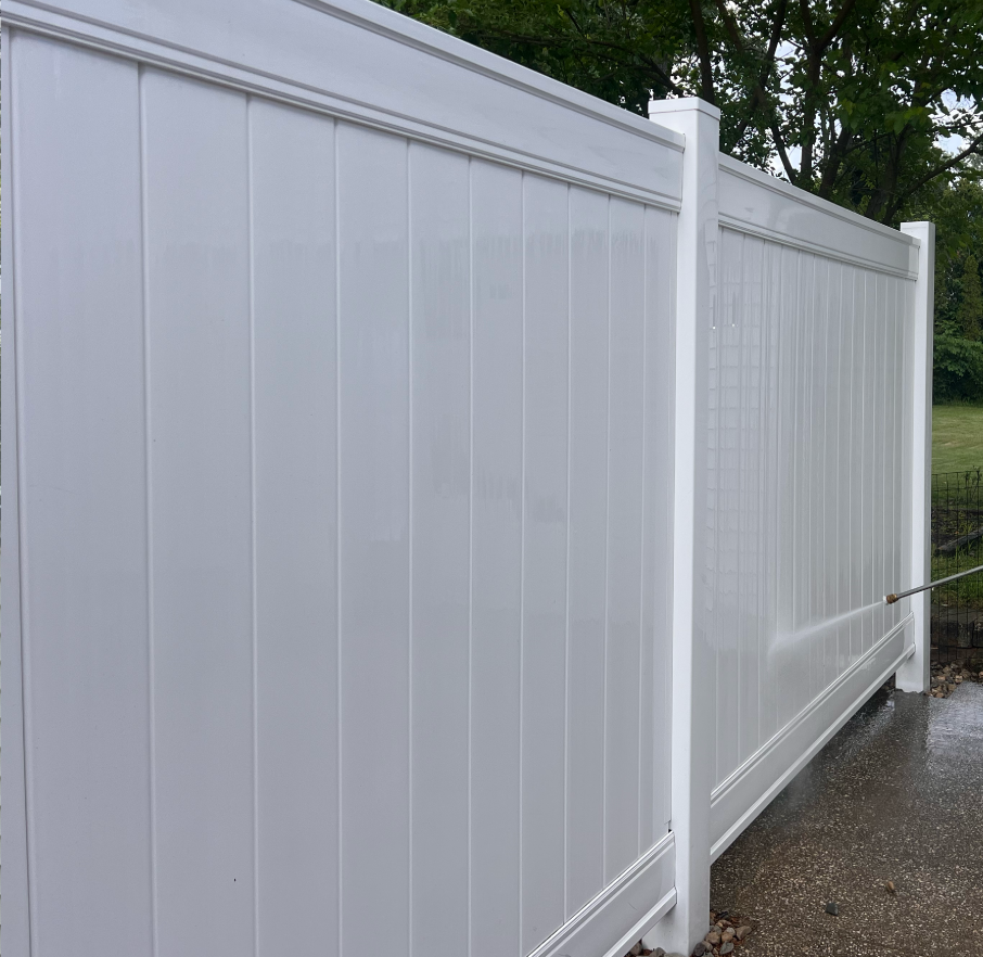 White vinyl privacy fence with vertical panels and decorative top trim, situated on a concrete surface with some green grass and trees in the background.