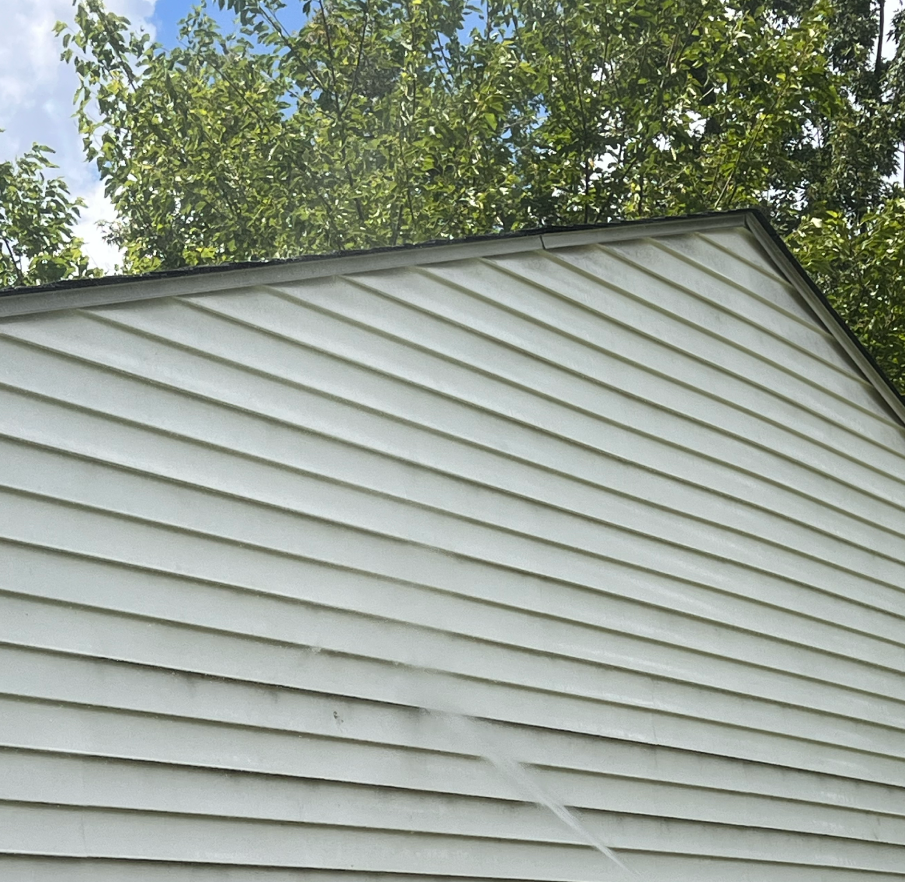 Close-up of the side of a house with white horizontal siding and a sloped roof, with a background of green trees and blue sky.