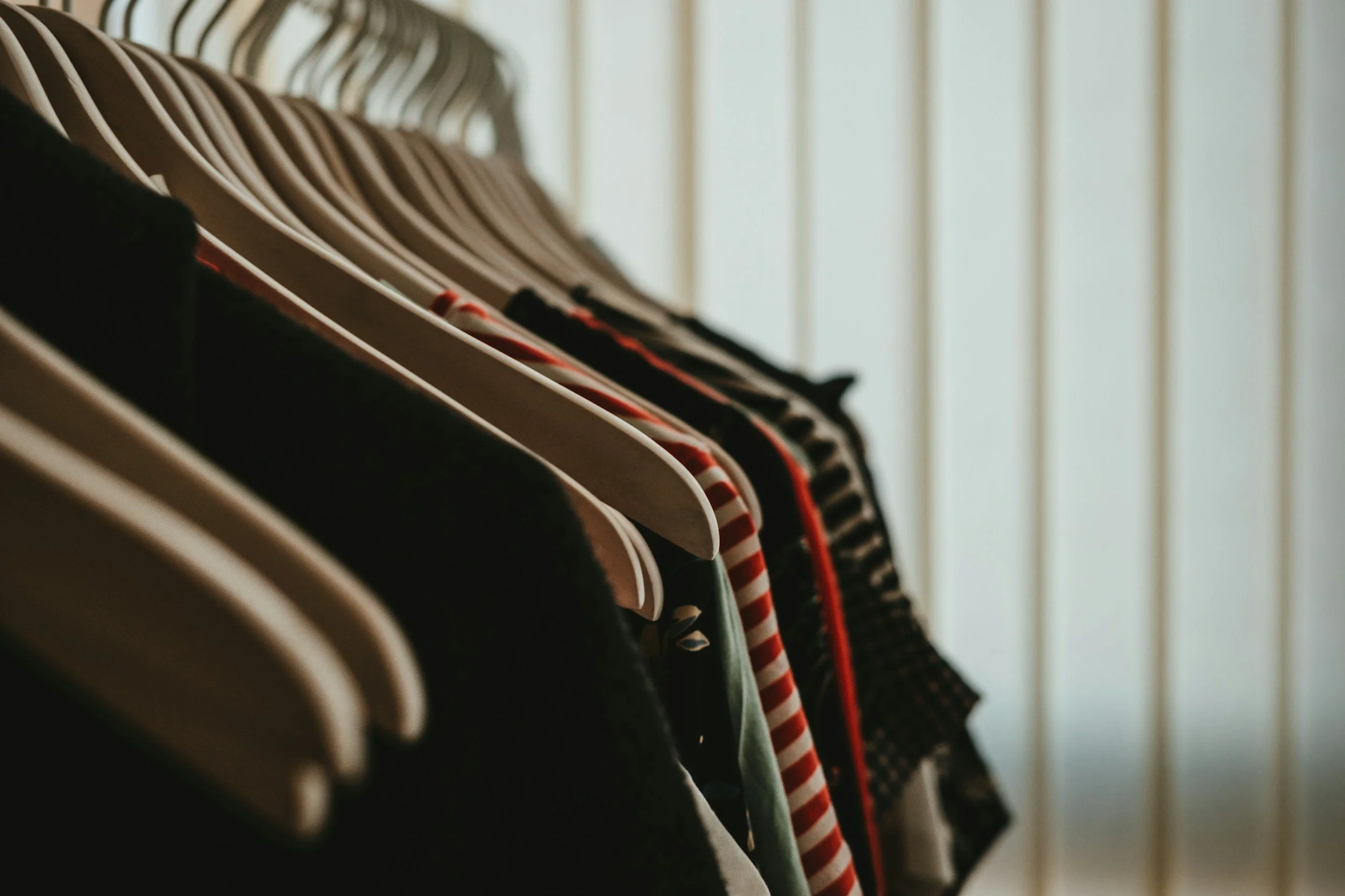 Clothes hanging on hangers in a closet, with sunlight coming through vertical blinds in the background.