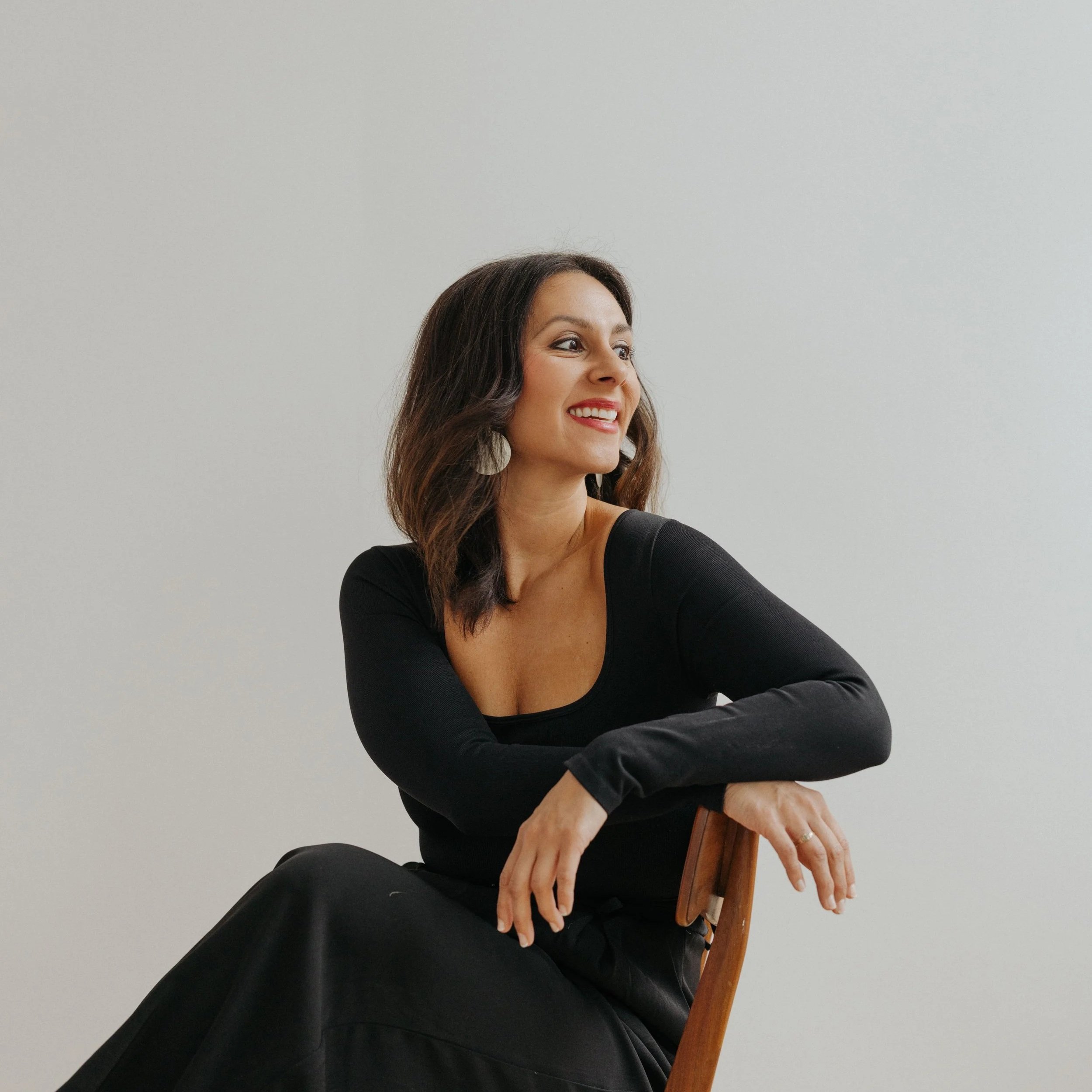 A woman with long brown hair smiling and looking to the side, sitting on a wooden chair, wearing a black top against a plain light grey background.