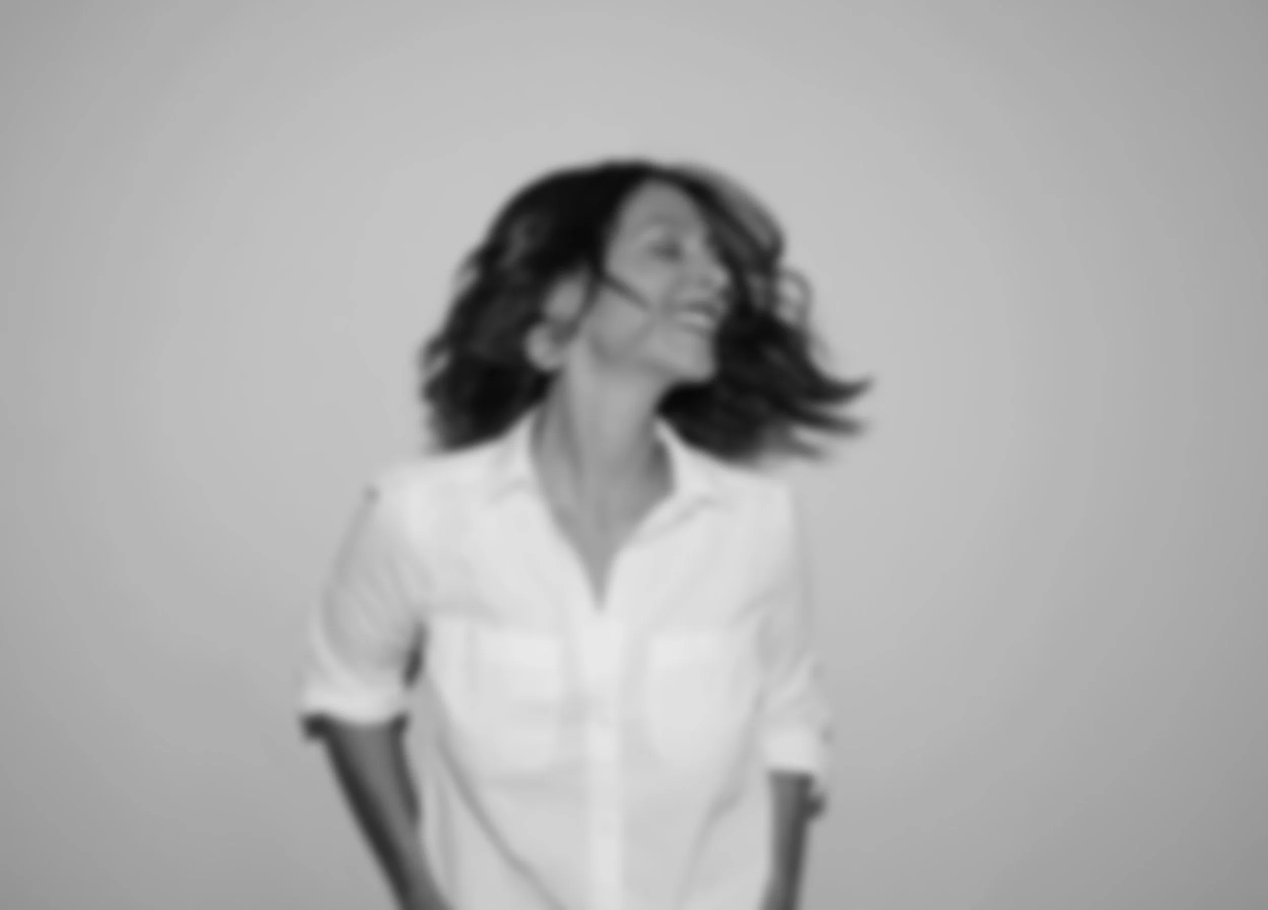 A woman with shoulder-length black hair smiling and wearing a white shirt, taken from below against a plain gray background.
