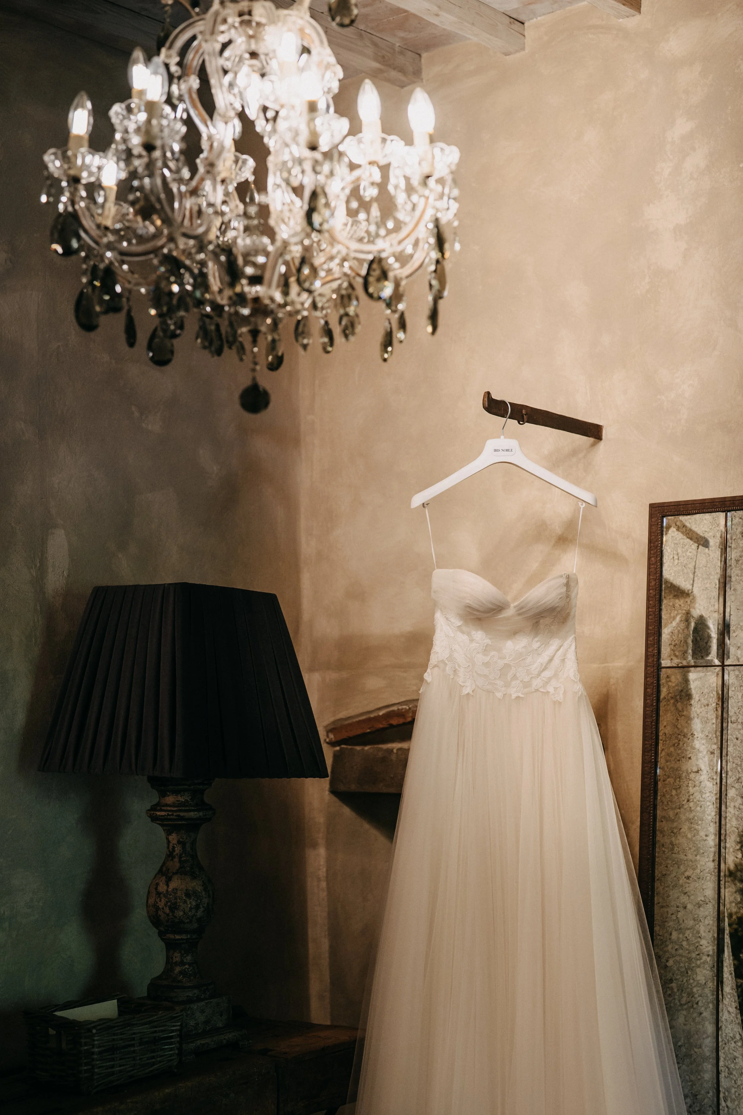 A white wedding dress with lace detailing hanging on a hanger with a name tag that reads 'IRIS NOBLE' in a rustic room. A large crystal chandelier hangs overhead, and a black lamp with a pleated shade is on a table below the dress. The room has textured beige walls and wood beams on the ceiling.