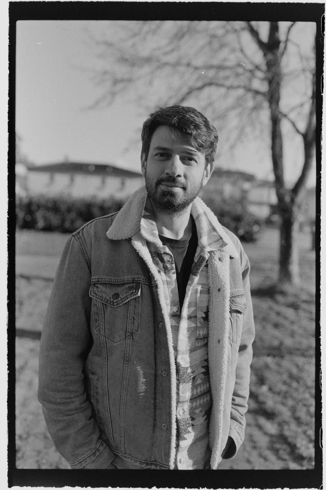 Black and white photo of a man with a beard and mustache, wearing a denim jacket with a sherpa lining, standing outdoors in front of leafless trees and a building.