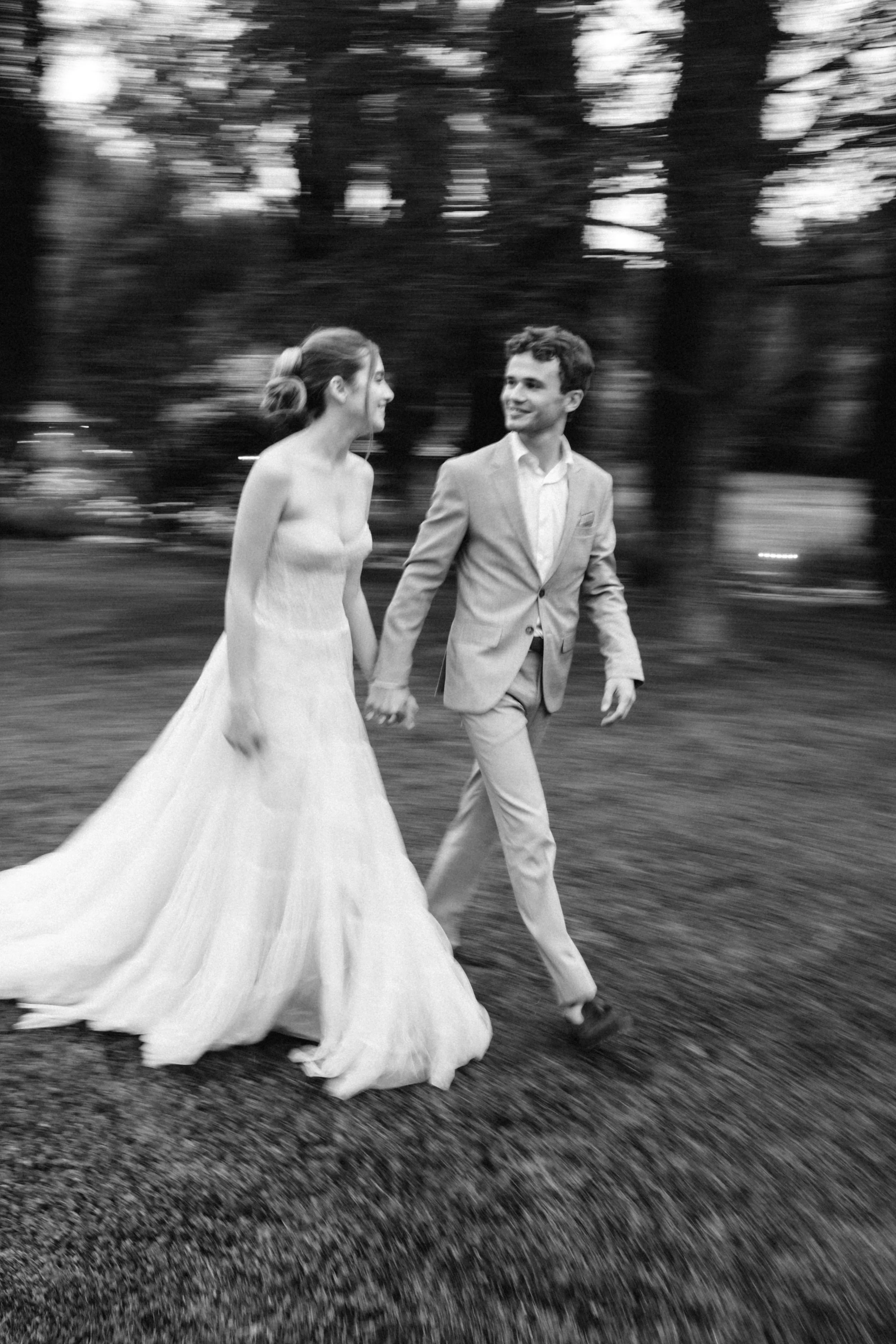 A black and white photo of a bride and groom walking hand in hand outdoors, with trees and a blurred background.