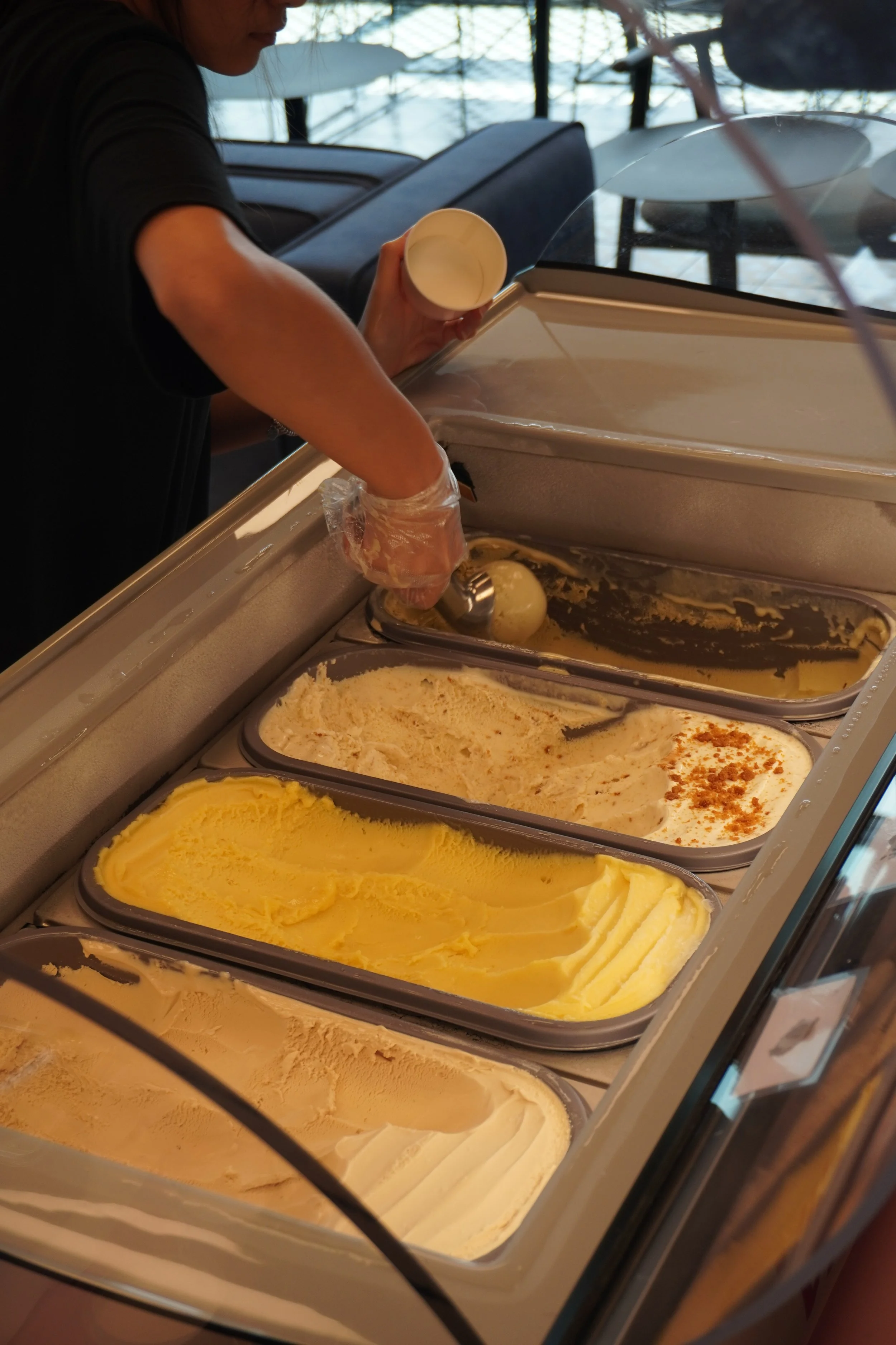 A person scooping ice cream from a tray at an ice cream shop, with multiple flavors visible in containers.