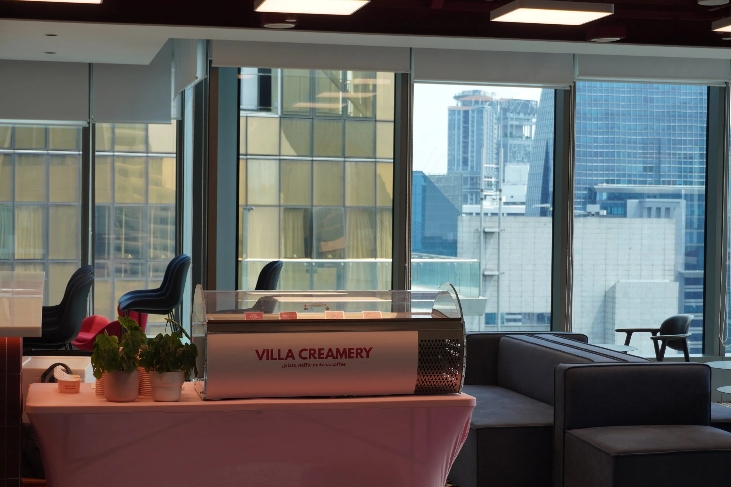 Interior of a modern office or café space with a view of city buildings through large windows. A table with potted plants and a dessert display labeled 'Villa Creamery' is in the foreground, with chairs and a seating area visible.