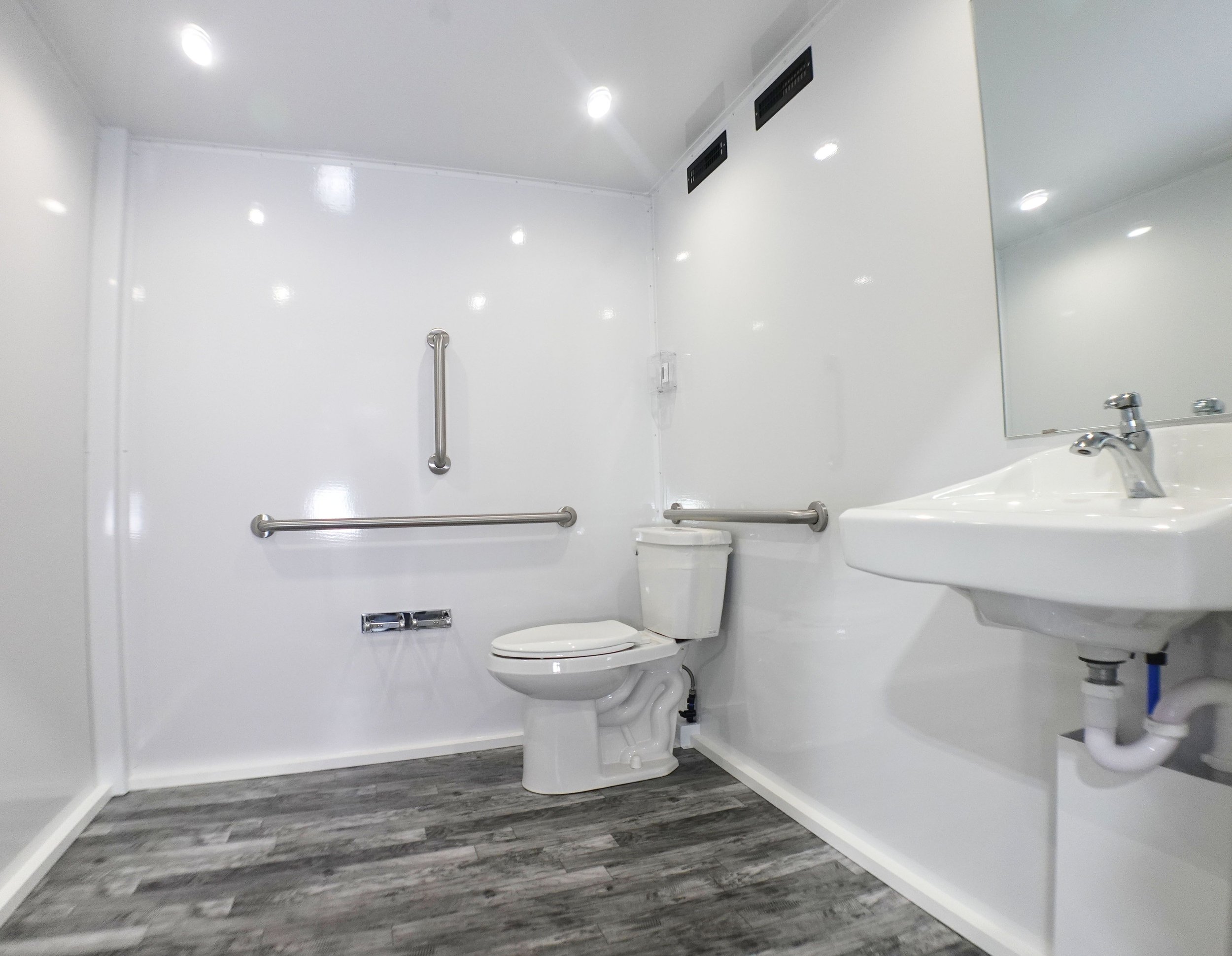 A clean, white bathroom featuring a toilet with grab bars, a sink with a faucet, and a large mirror. The ceiling has recessed lighting and air vents. The floor has wood-like gray tiles.