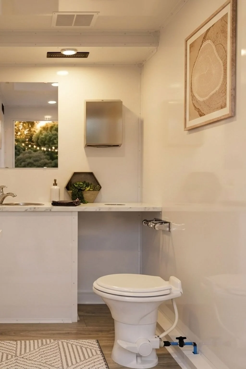 A bathroom with a white toilet, a white marble countertop, and a small sink. There is a hexagonal shelf with plants and a soap dispenser. A framed artwork hangs on the wall, and a window shows trees outside. A mirror and ceiling lights are also visible.