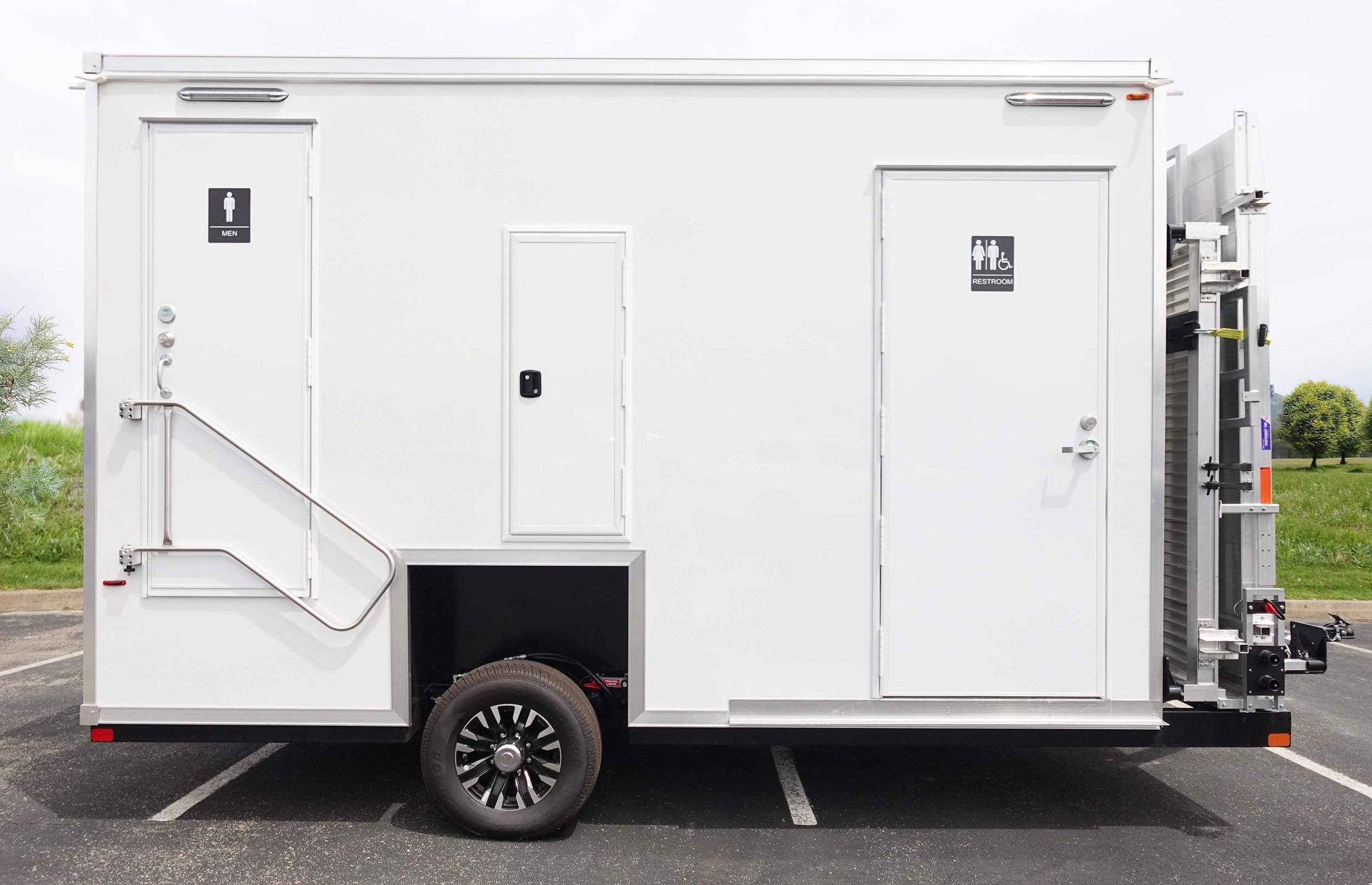 A white mobile restroom trailer with two doors marked for men and women, situated in a parking lot with a grassy area and trees in the background.