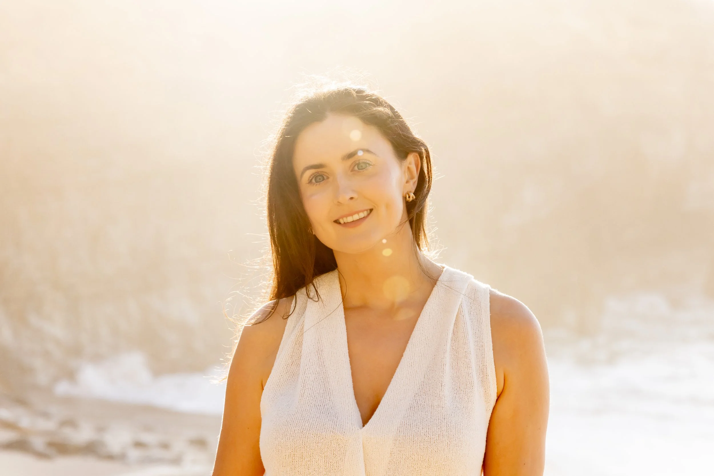 A woman with brown hair and green eyes smiling at the camera, standing outdoors in bright sunlight during sunset or sunrise, with a blurred background of water and a sunlit sky.