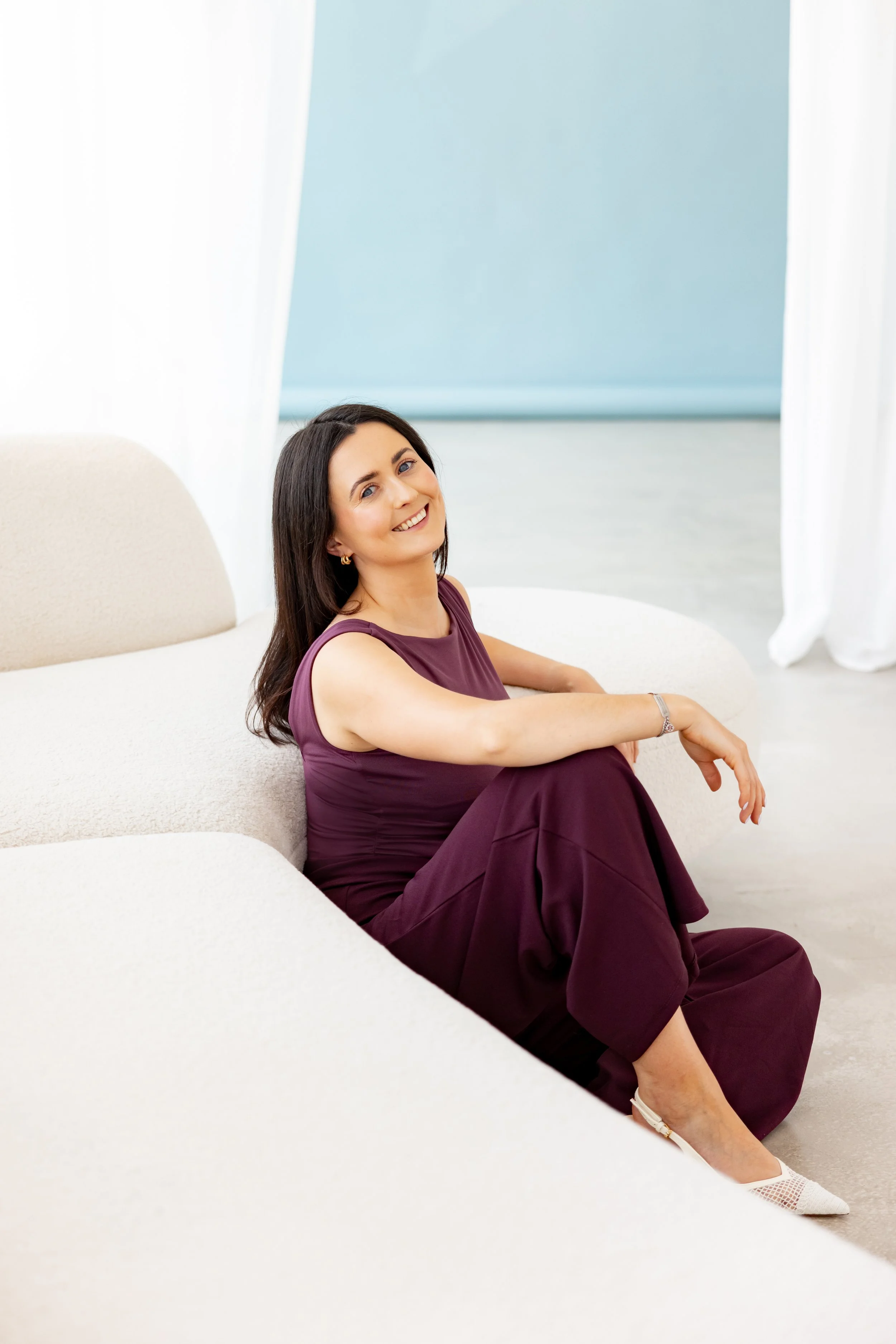 A woman with dark hair, wearing a sleeveless purple dress and white shoes, sitting on a white couch in a bright, minimalist room with light blue walls and white curtains, smiling at the camera.