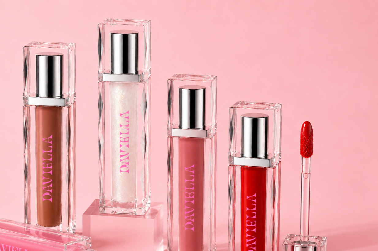 Four lip glosses in clear glass bottles with metallic caps, labeled 'DAVADEA,' with a pink and red color scheme, one with a doe-foot applicator, arranged on pink background.