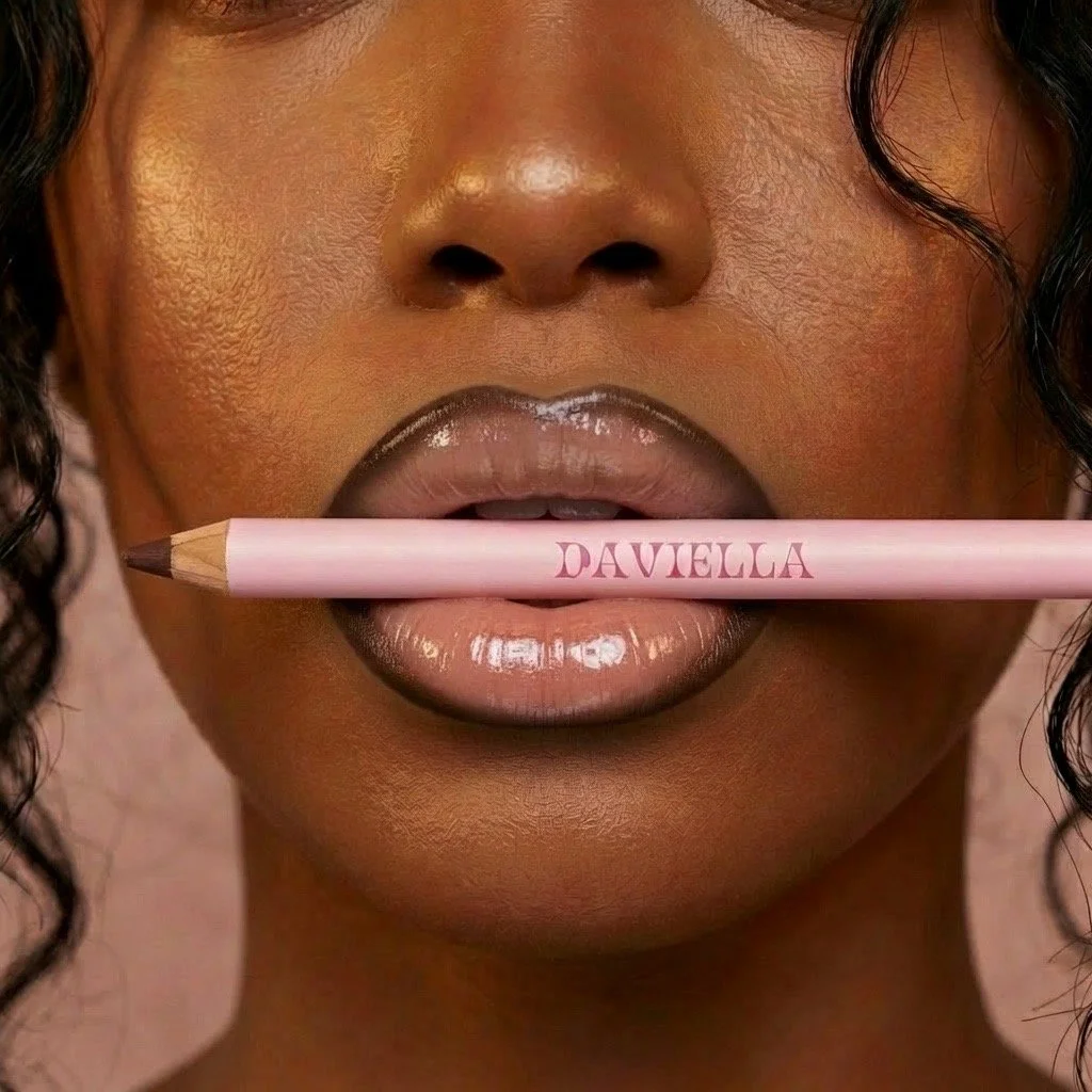 Close-up of a woman with dark curly hair holding a pink pencils with the name 'DAVIELLA' in her lips.