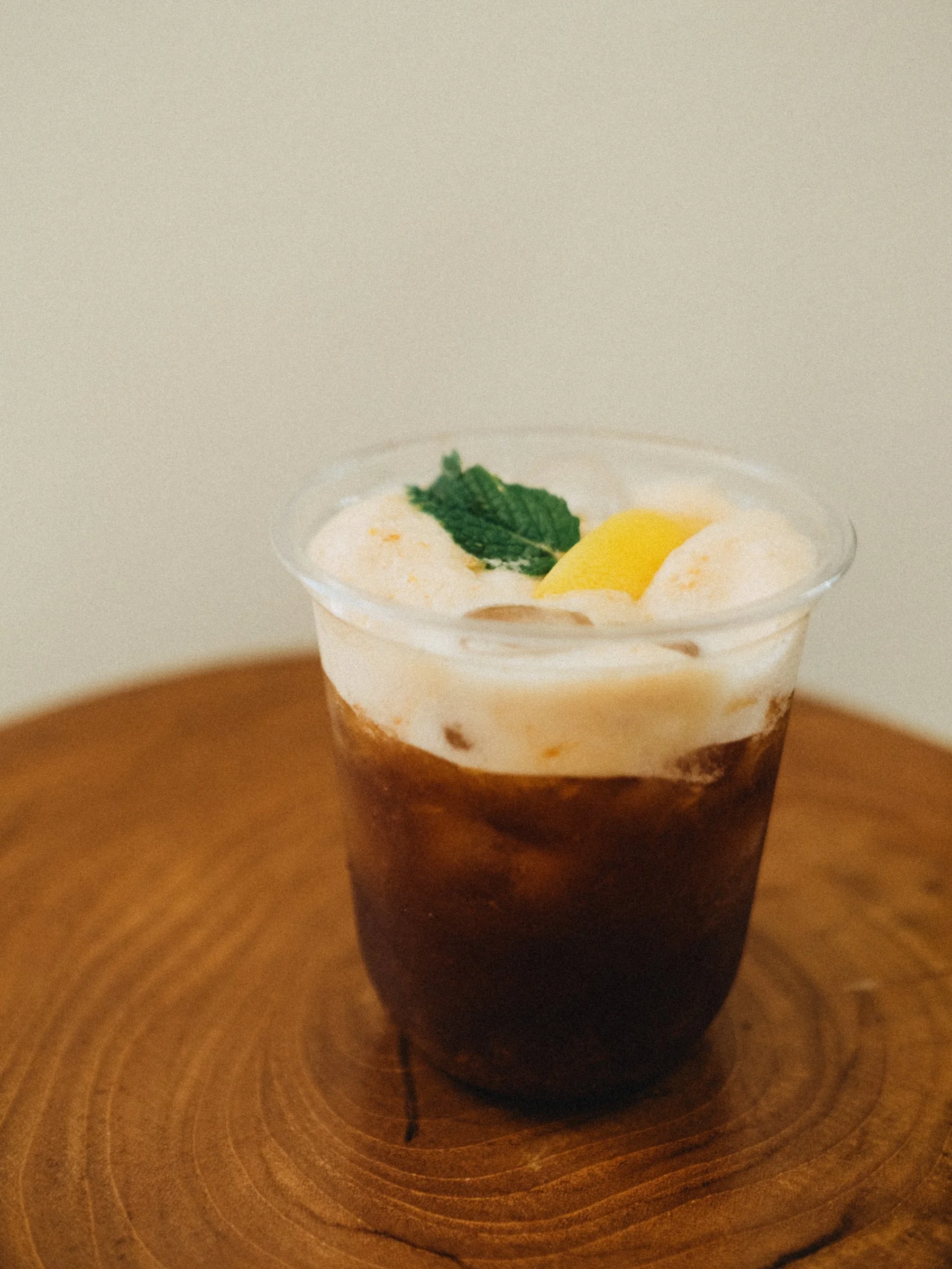 A clear plastic cup of peach honey shaken with cold brew with a mint leaf and lemon slice on top, served on a wooden table.