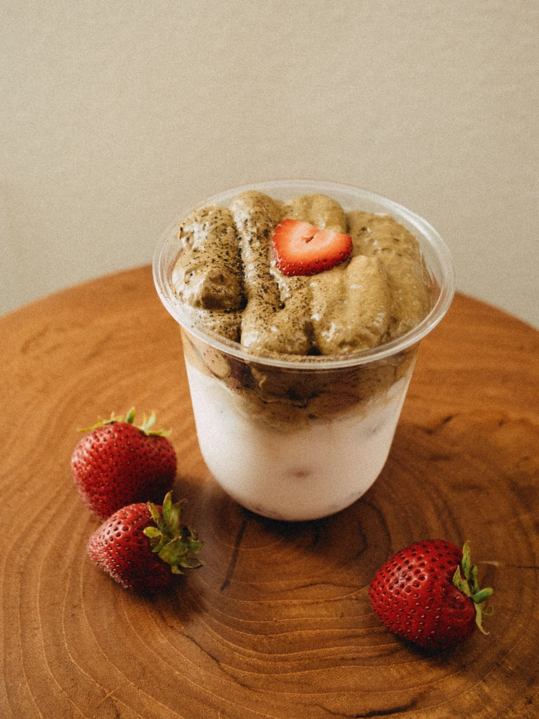 A clear plastic cup of strawberry honey milk topped with a sliced strawberry and a sprinkle of hojicha powder, placed on a wooden surface with whole strawberries nearby.