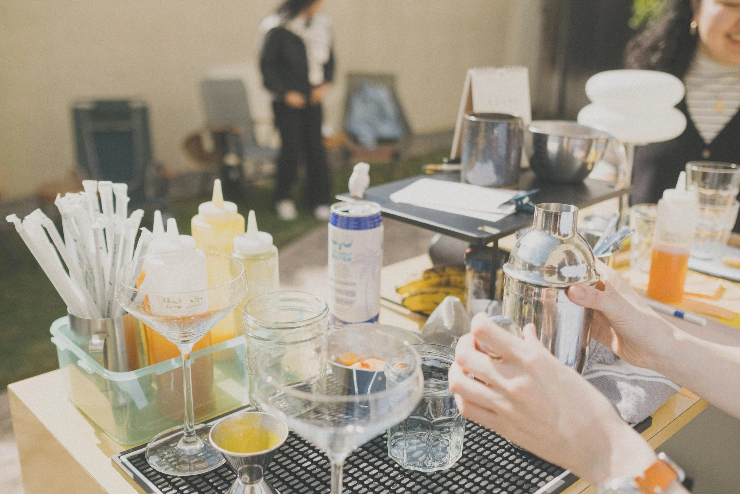 A person preparing drinks at an outdoor bar or event station, with various bottles, mixing tools, and ingredients on the counter, including a cocktail glass, whipped cream, and bottles of flavored syrups.