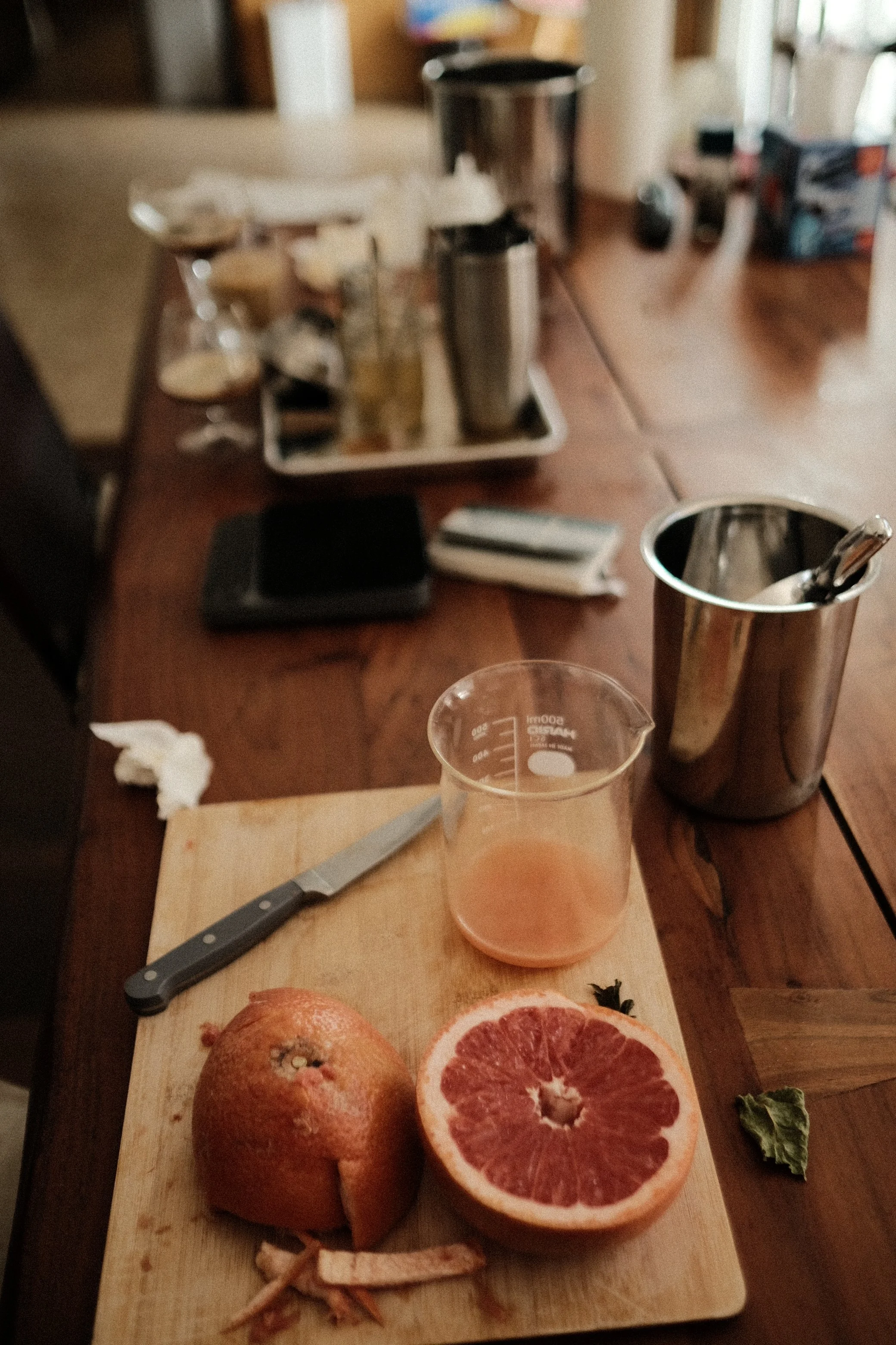 A cutting board with a partially peeled grapefruit, a halved grapefruit, and a whole grapefruit. There is also a small herb and a knife on the cutting board. In the background, a glass with a pink beverage, a metal tumbler, and a tray with various it