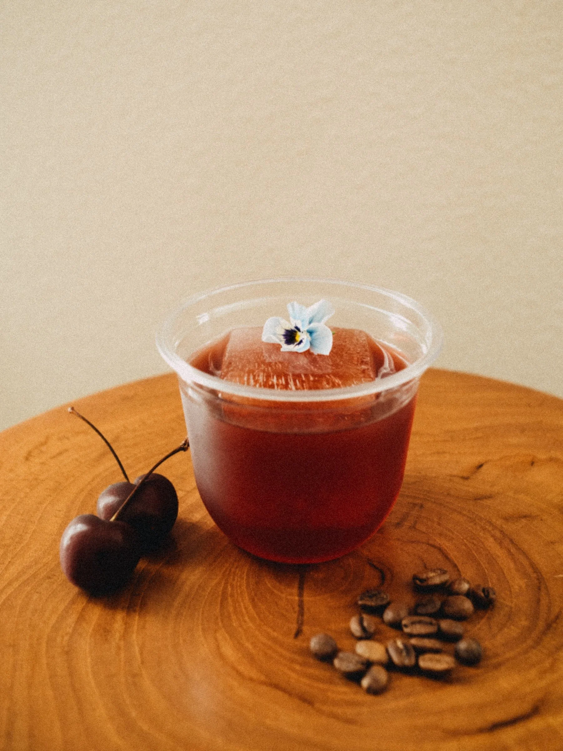 A glass of iced clarified cherry coffee with a violet flower garnish on top, placed on a wooden surface with cherries and coffee beans beside it.